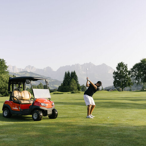 Ein Golfer in schwarzem Shirt und weißen Shorts schwingt seinen Schläger auf einem grünen Golfplatz mit Bergen im Hintergrund.