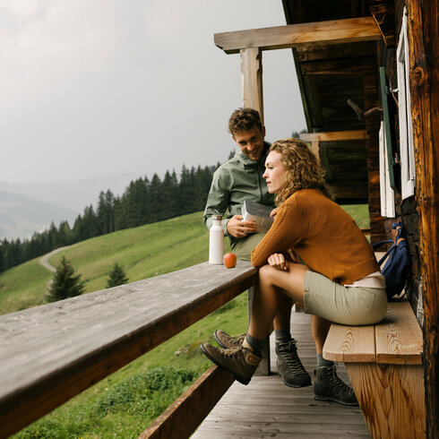 Wandern Einkehr Kitzbuehel Sommer Huette (c) Kitzbuehel Tourismus (24) Zwei Personen sitzen vor einer Berghütte.