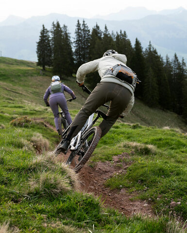 Zwei Mountainbiker fahren auf einem schmalen Pfad durch eine grüne Wiese, umgeben von hohen Bäumen und Bergen im Hintergrund.